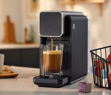 Schwarze »Qbo ONE« Kapselmaschine, Soft Black, die Kaffee in ein Glas füllt. Daneben ein Korb mit Kaffeekapseln.
