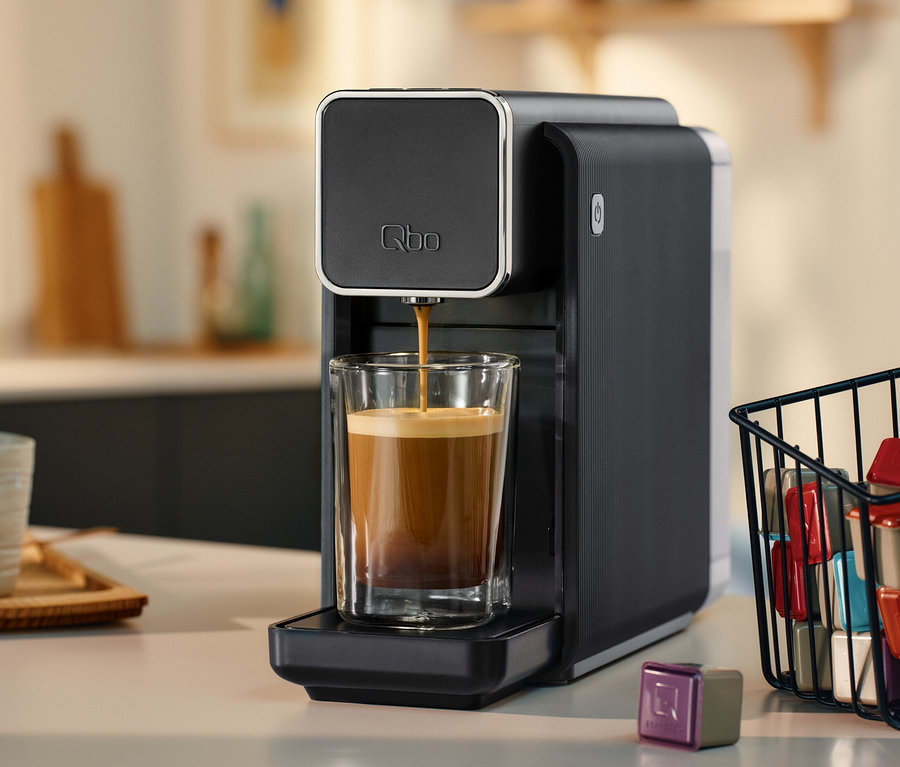 Schwarze »Qbo ONE« Kapselmaschine, Soft Black, die Kaffee in ein Glas füllt. Daneben ein Korb mit Kaffeekapseln.