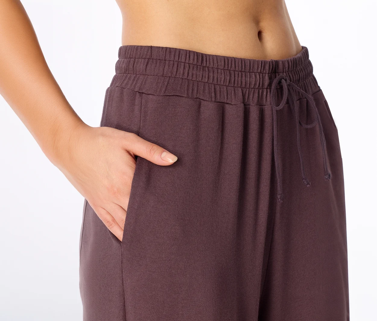 Nahaufnahme einer Person, die eine Hand in die Tasche einer dunkelvioletten Jogginghose mit Gummibund und Kordelzug steckt.