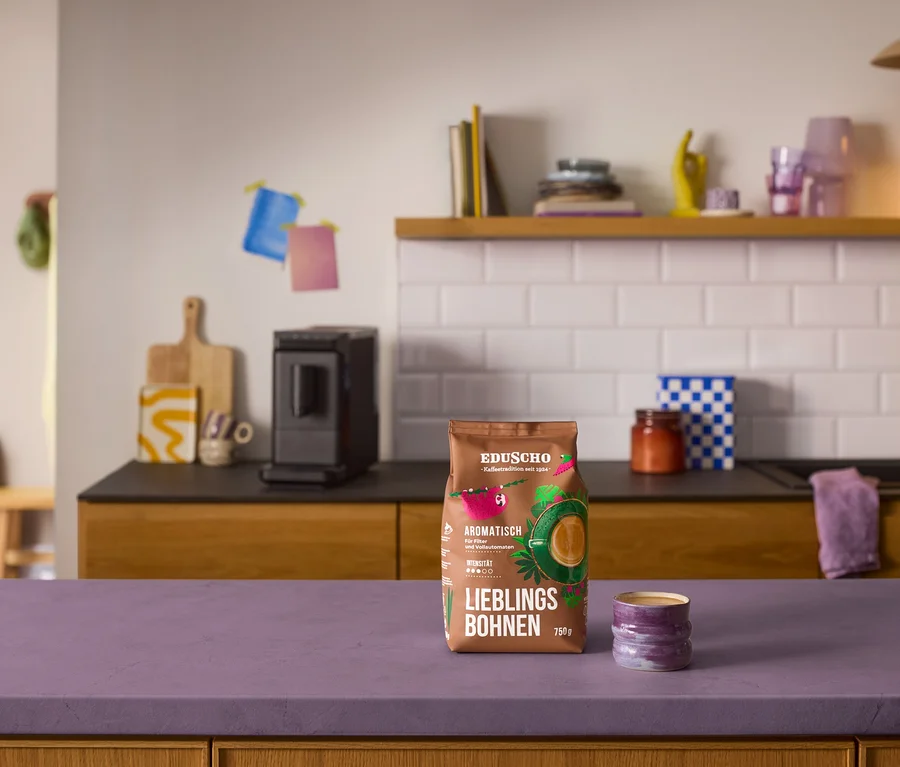 Ein brauner Beutel Eduscho Lieblingsbohnen Aromatisch und eine kleine lila Tasse stehen auf einer lila Arbeitsplatte. Im Hintergrund ist ein schwarzer »Esperto mini« Kaffeevollautomat auf einer Küchenzeile mit weißen Fliesen zu sehen.