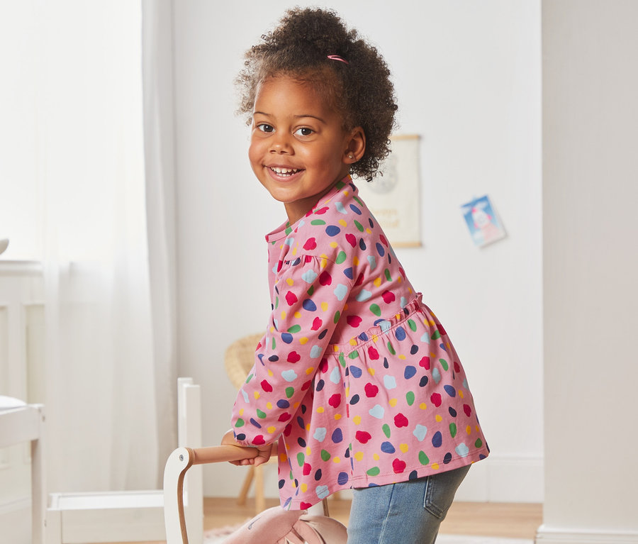 Lächelndes Mädchen in einem rosa Langarmshirt aus Bio-Baumwolle (2er-Pack) steht in einem Kinderzimmer.