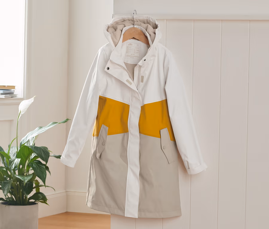 Ein weißer, gelber und beiger Regenparka hängt auf einem Kleiderbügel an einer weißen Wand.