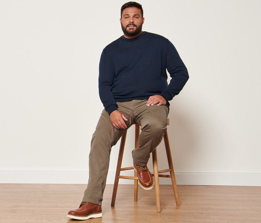 Mann sitzt auf einem Hocker, trägt marineblauen Merinopullover und sandfarbene Chino.