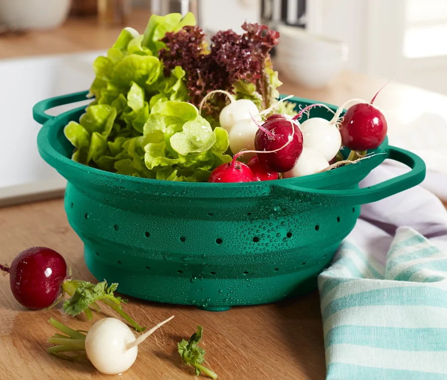 Ein grünes Platzspar-Sieb voll mit Salat und Radieschen steht auf einem Holztisch.