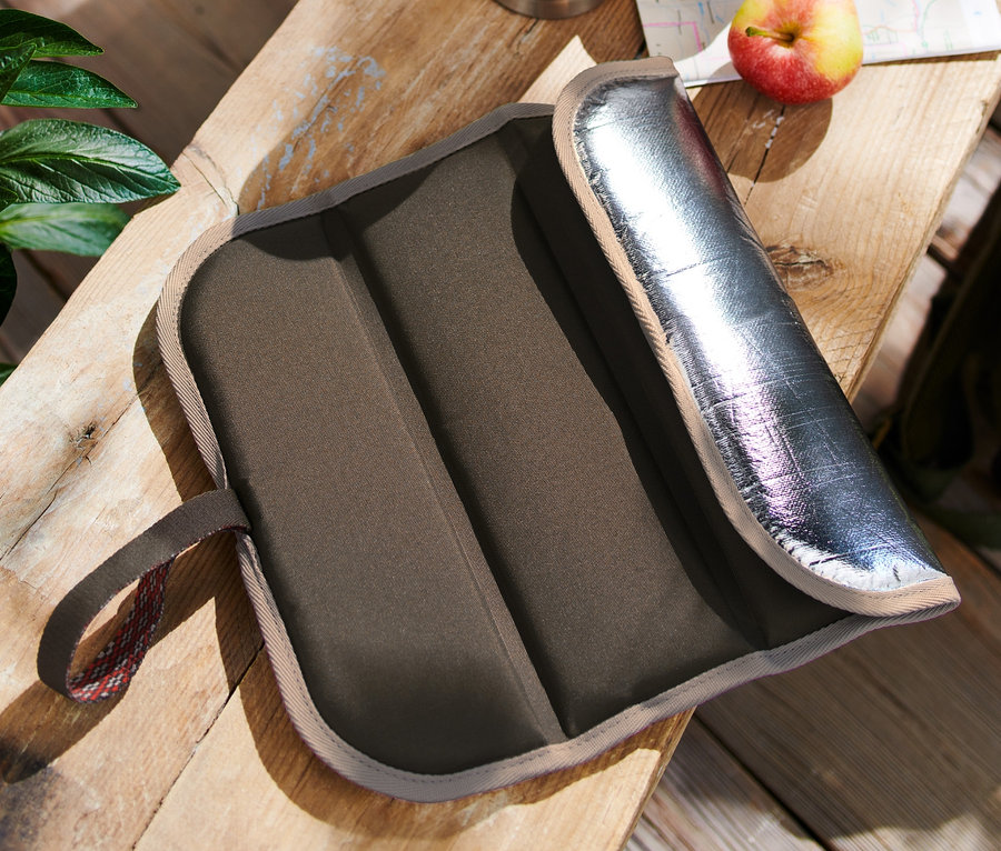 Ein faltbares Thermo-Sitzkissen liegt auf einem Holztisch neben einer Thermoskanne, einem Apfel und einem Rucksack.