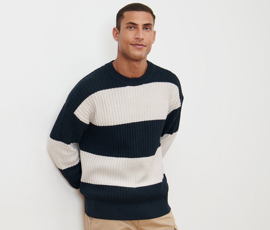 Mann posiert vor weißem Hintergrund mit einem gestreiften Grobstrick-Pullover in Marineblau und Weiß und Khaki-Hosen.