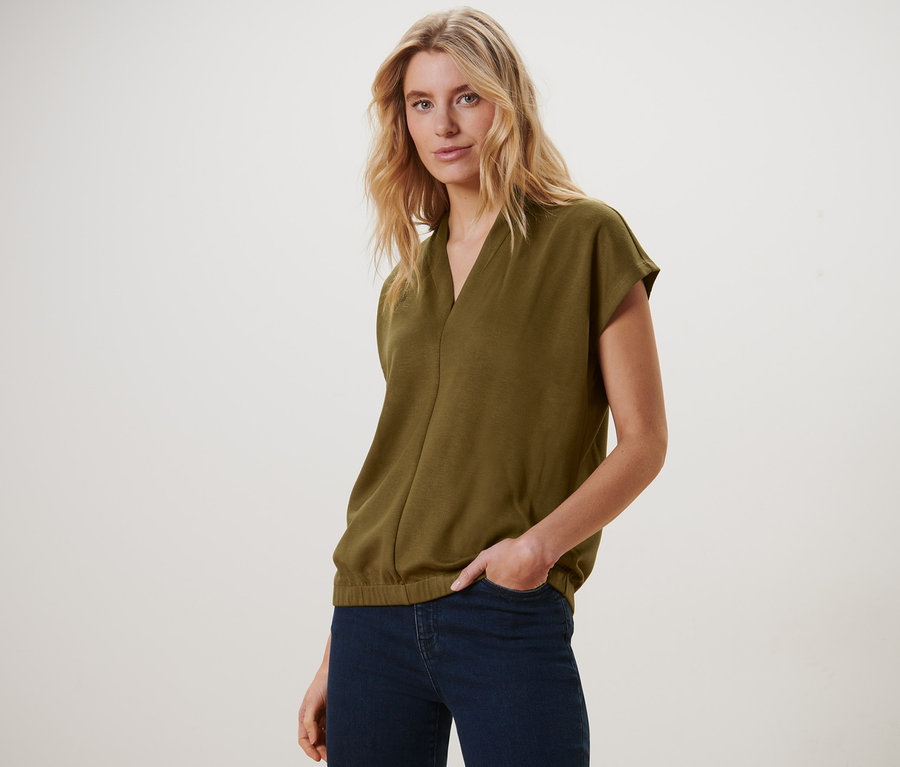 Blonde Frau posiert in einem olivgrünen Blusenshirt mit V-Ausschnitt und dunkelblauen Jeans.