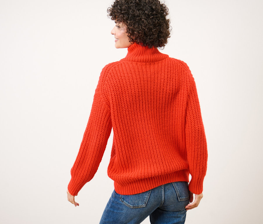 Rückansicht einer Frau mit lockigem Haar, die einen orangefarbenen Grobstrick-Troyer und blaue Jeans trägt.