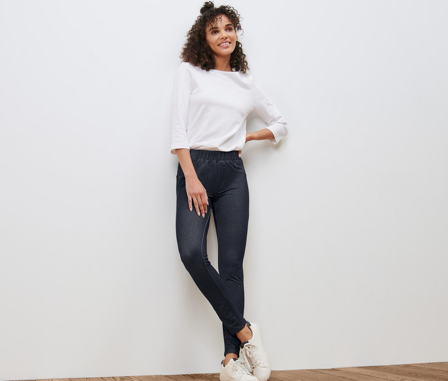 Frau, die an einer weißen Wand lehnt, trägt Leggings in Jeansoptik, eine weiße Bluse und Turnschuhe.