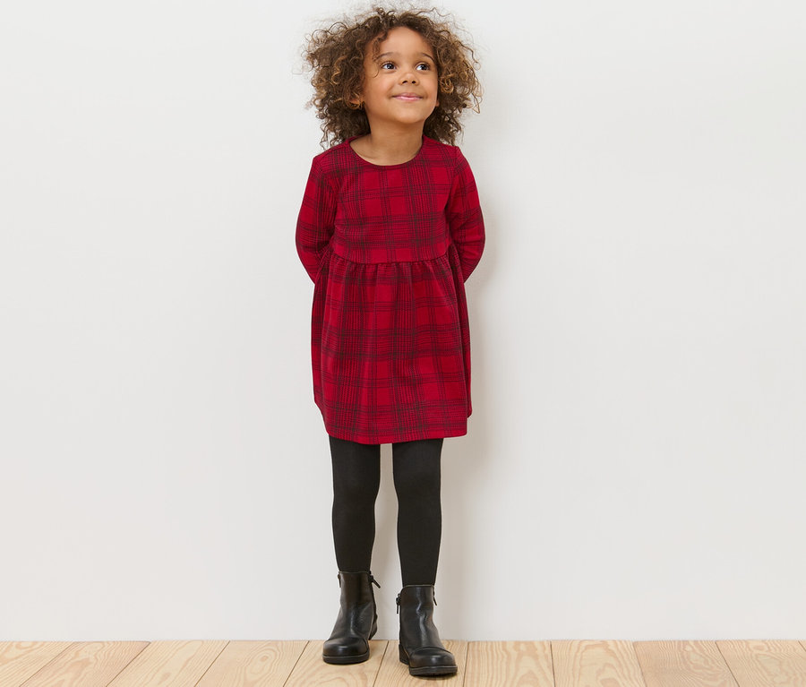 Ein Mädchen steht mit den Händen hinter dem Rücken und trägt ein Kinder-Jersey-Kleid, eine schwarze Leggings und schwarze Schuhe.