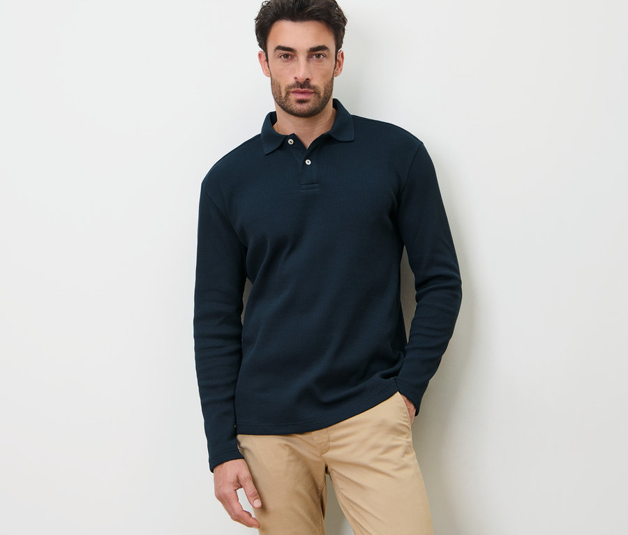 Ein Mann posiert vor einem weißen Hintergrund in einem dunkelblauen Langarm-Poloshirt.