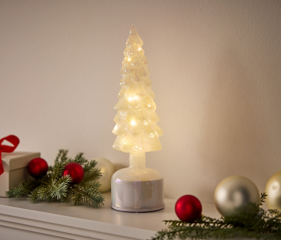 Dekorativer beleuchteter Baum steht neben Geschenk und Kugeln auf einer weißen Oberfläche.