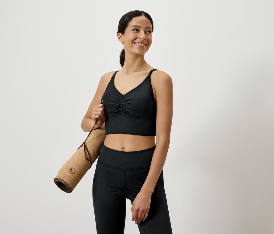 Lächelnde Frau in schwarzer Sportbekleidung hält eine zusammengerollte Yogamatte unter dem Arm.