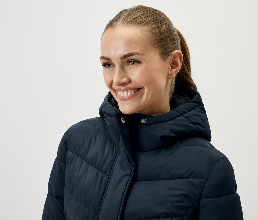 Frau mit braunen, zum Pferdeschwanz gebundenen Haaren lächelt und trägt eine dunkelblaue Daunenjacke.