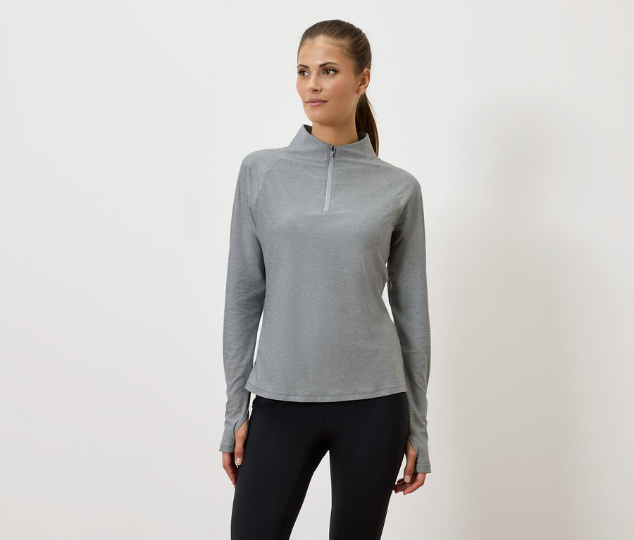 Frau in grauem Reißverschluss-Sweatshirt und schwarzen Leggings steht vor einem weißen Hintergrund.