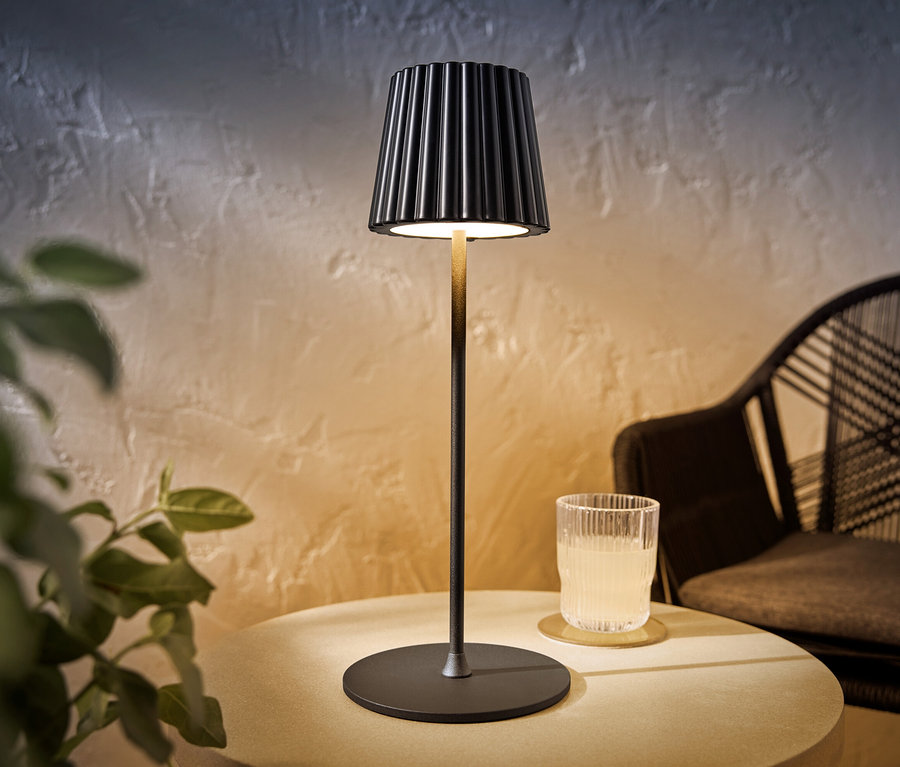 Eine schwarze LED-Akku-Outdoor-Tischleuchte steht auf einem Tisch neben einem Glas mit einem Getränk.