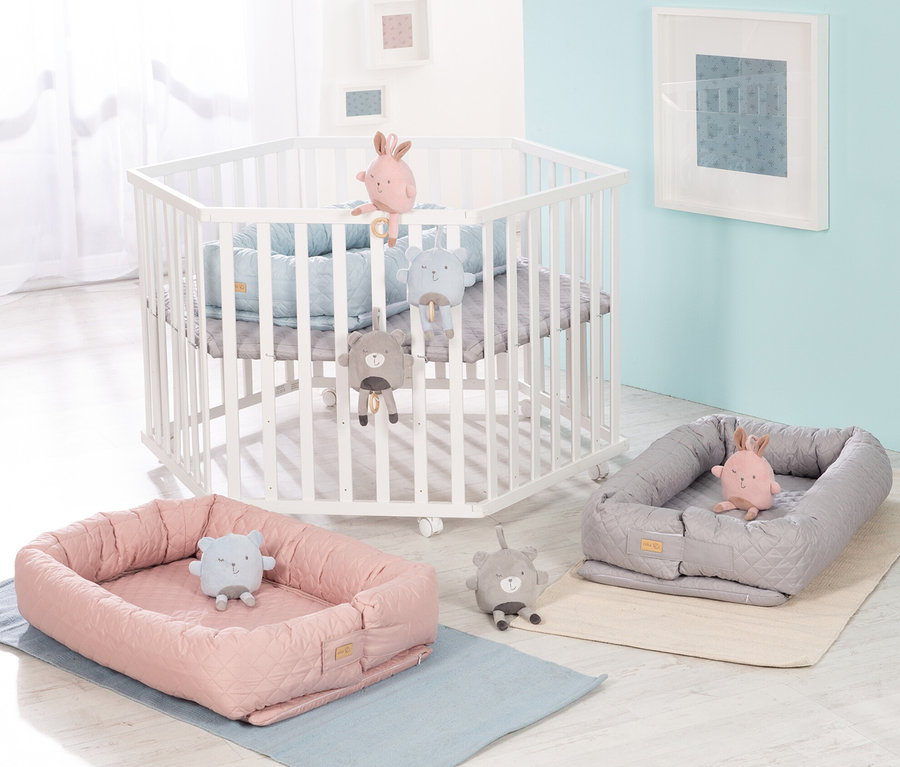 Weißes Kinderbett mit grauer Spieldecke, rosa und graue Babynester mit Plüschtieren auf Unterlagen.