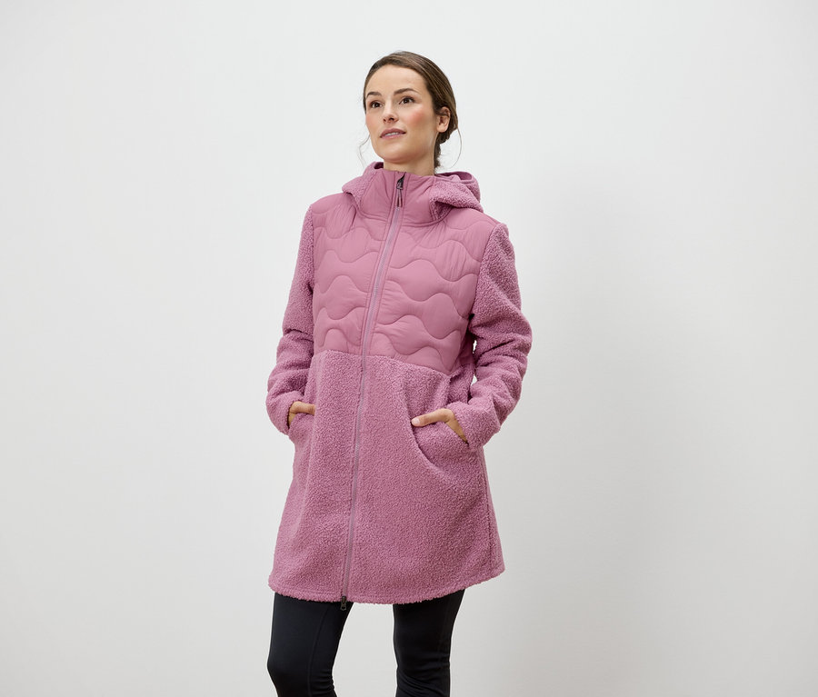 Frau steht mit den Händen in den Taschen eines rosafarbenen Reissverschlussmantels mit Kapuze.