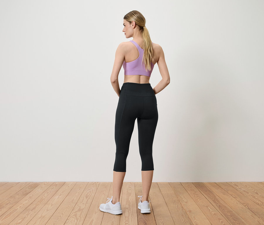 Frau posiert mit dem Rücken zur Kamera und trägt einen lila Sport-BH, schwarze Leggings und weiße Turnschuhe.