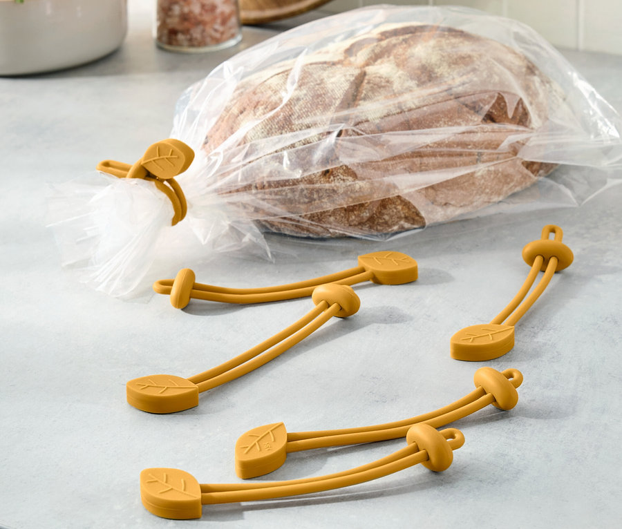 Brot in Plastiktüte ist mit einem gelben Gummiband mit Blattmotiv verschlossen. Drei weitere Gummibänder liegen daneben.