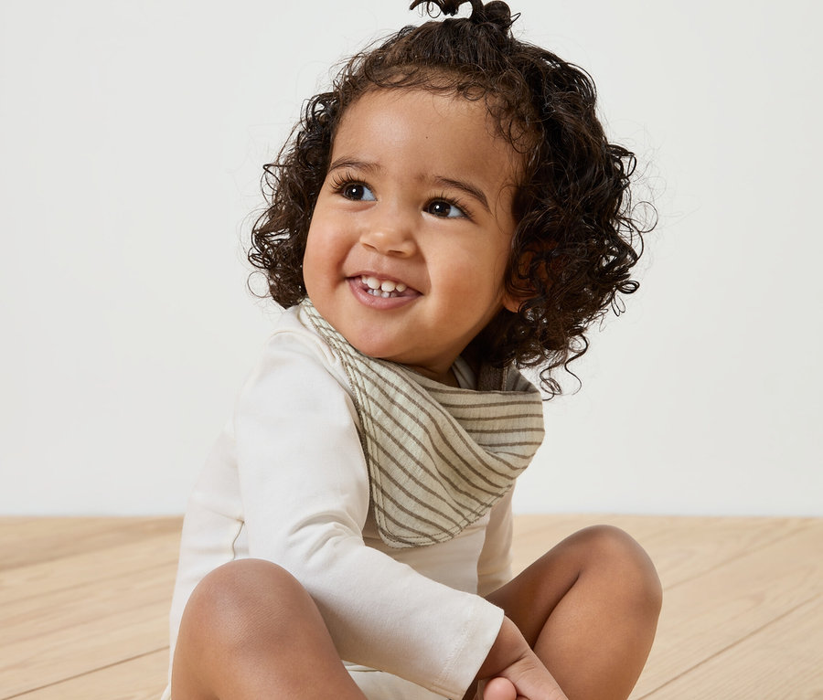 Lächelndes Baby sitzt auf Holzboden und trägt ein 5er-Pack Baby-Musselin-Halstücher.