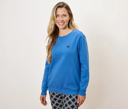 Eine Frau trägt ein blaues Langarmshirt mit einem kleinen schwarzen Herz und gemusterte Hosen.