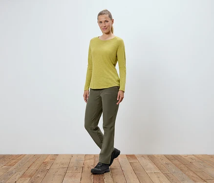 Eine Frau in olivgrüner Hose, gelbgrünem Langarmshirt und schwarzen Schuhen steht auf einem Holzboden vor einer weißen Wand.