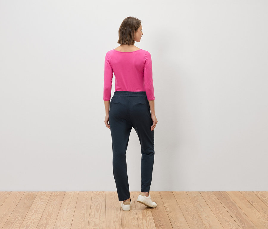 Eine Frau steht mit dem Rücken zur Kamera, sie trägt eine pinkfarbene Bluse und blaue Hosen.