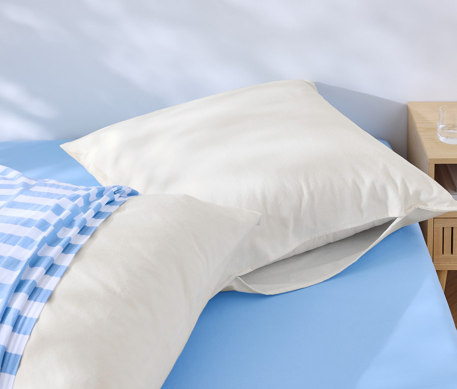 Zwei weiße Kopfkissen liegen auf einem blauen Bett, eines davon mit einem blau-weiß gestreiften Bezug. Neben dem Bett steht ein Nachttisch mit Wasser und einem Telefon.