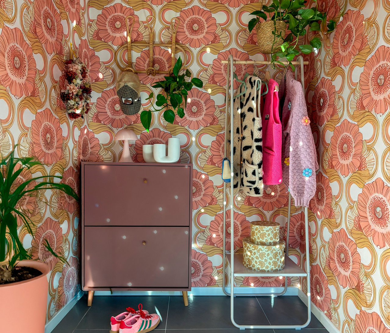 Ein Schuhschrank mit zwei Schubladen steht vor einer geblümten Tapete. An einer Kleiderstange hängen eine Weste mit Tiermuster und zwei weitere Westen. Auf dem Boden stehen rosa Turnschuhe.