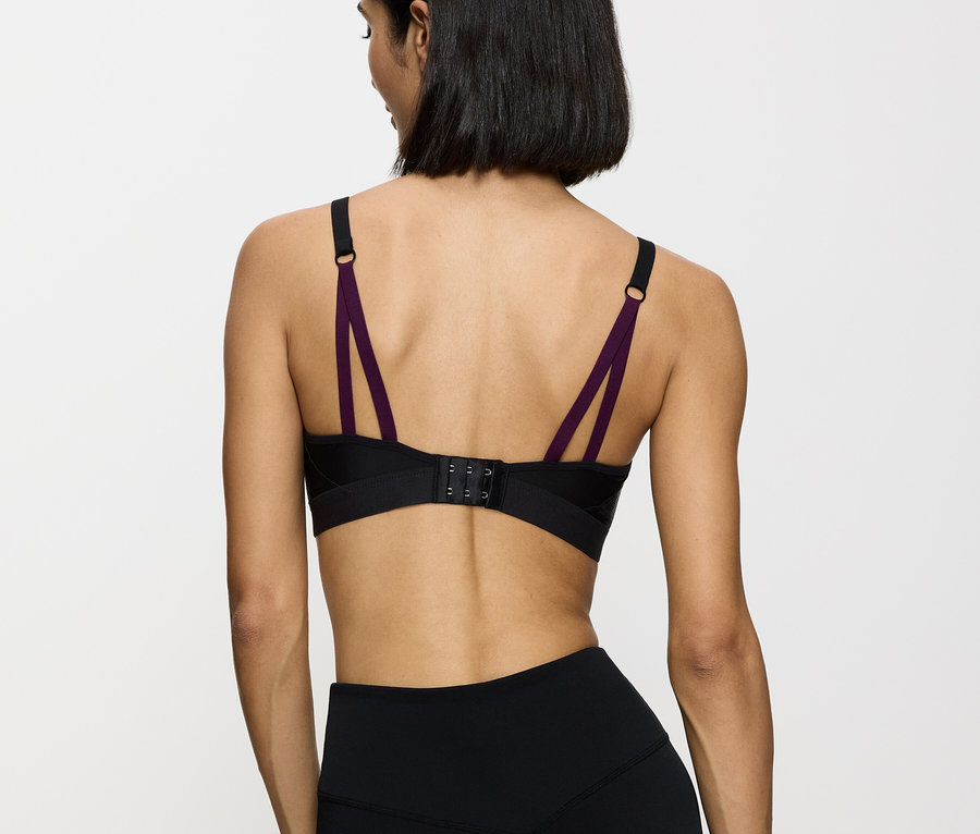 Rückansicht einer Frau mit schwarzem Sport-BH mit lila Trägern und schwarzen Leggings vor einem weißen Hintergrund.