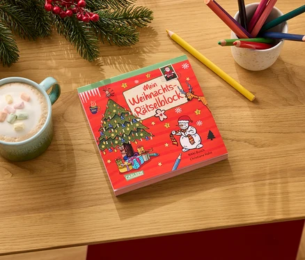 Auf einem Holztisch liegen ein roter Weihnachtsrätselblock, eine Tasse Kakao mit Marshmallows, Buntstifte und Tannenzweige.