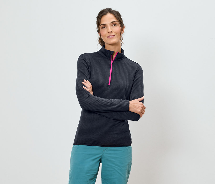 Frau mit verschränkten Armen trägt ein dunkelblaues, langärmeliges Hemd mit einem pinkfarbenen Reissverschluss und türkisfarbene Hosen vor einem weissen Hintergrund.