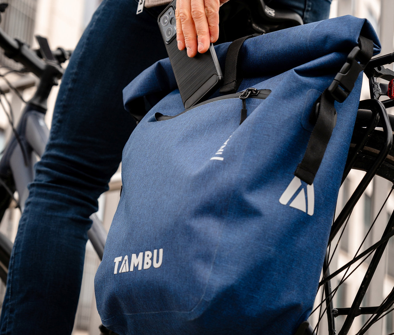 Eine Person verstaut ein Smartphone in einer blauen Tambu »INAYA« Fahrradtasche, die an einem Fahrrad befestigt ist.