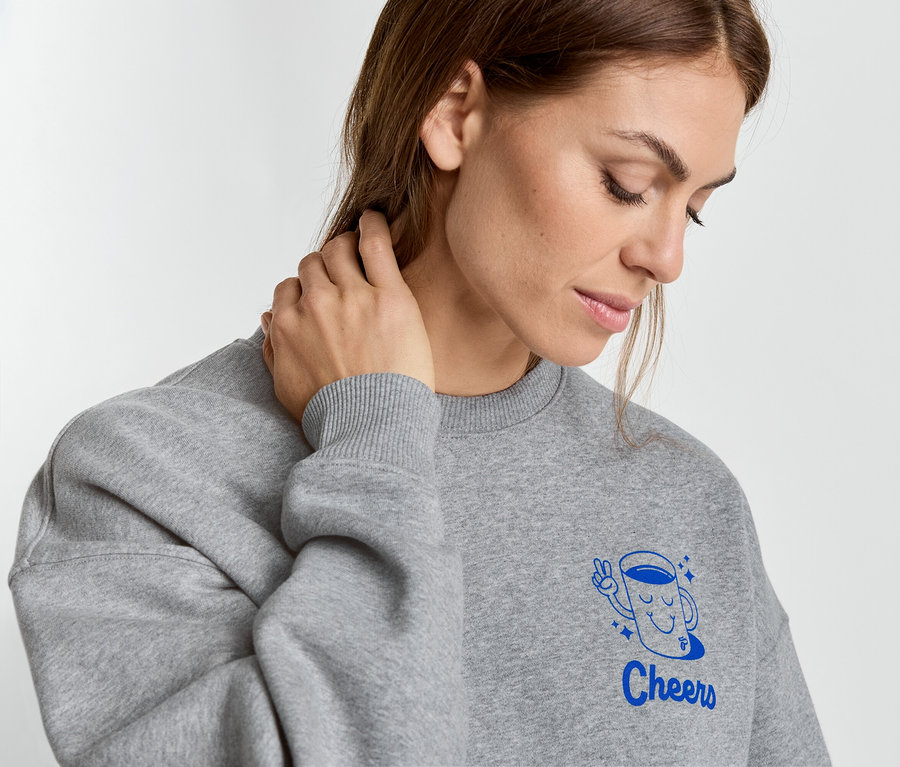 Frau mit grauem Pullover, der eine blaue Tasse mit der Aufschrift "Cheers" zeigt, berührt ihren Nacken.