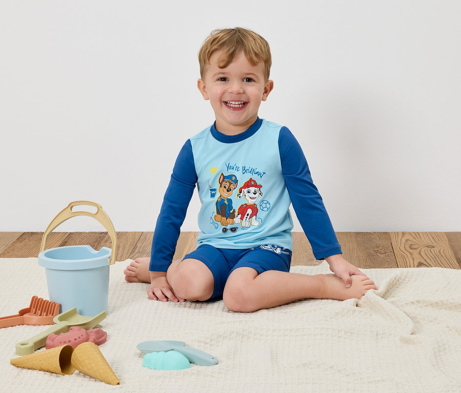 Ein lächelnder Junge sitzt auf einer Decke mit einem hellblauen Paw Patrol-Shirt und blauen Shorts, umgeben von Sandspielzeug.