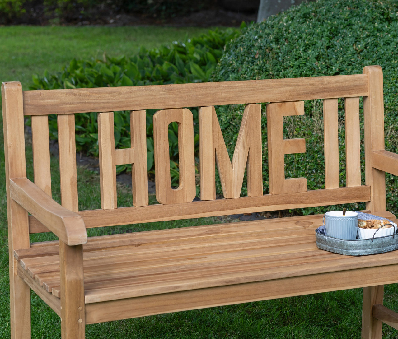 deVries Gartenbank »Home« steht auf einer Wiese, darauf ein Getränk mit Gebäck.