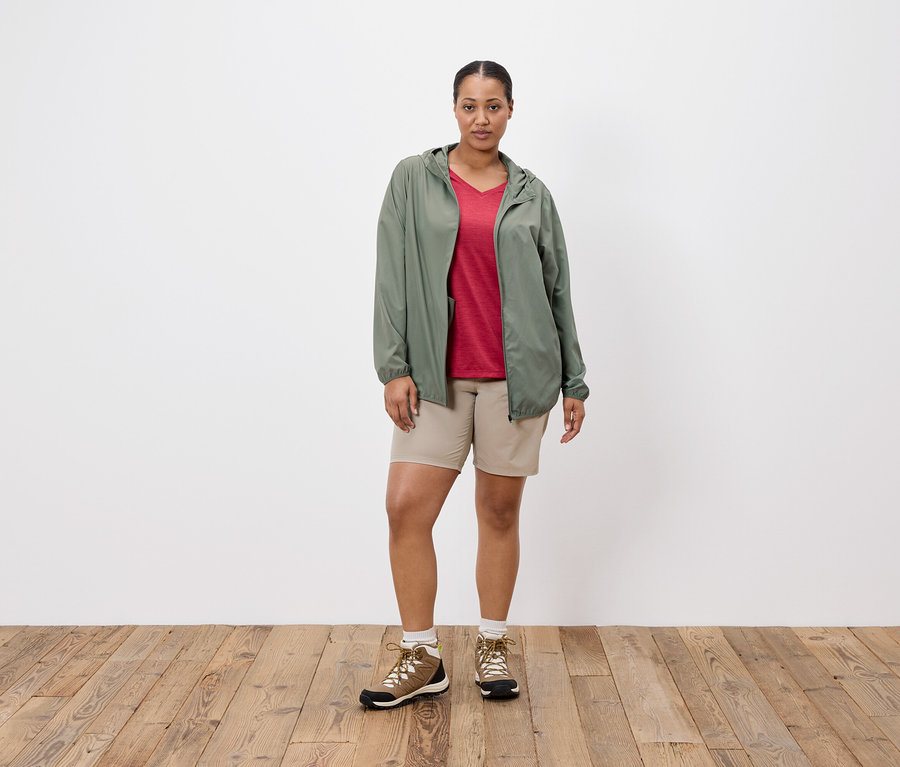 Frau steht vor einer weißen Wand, trägt eine grüne Jacke, ein rotes T-Shirt, beige Shorts und Wanderschuhe.