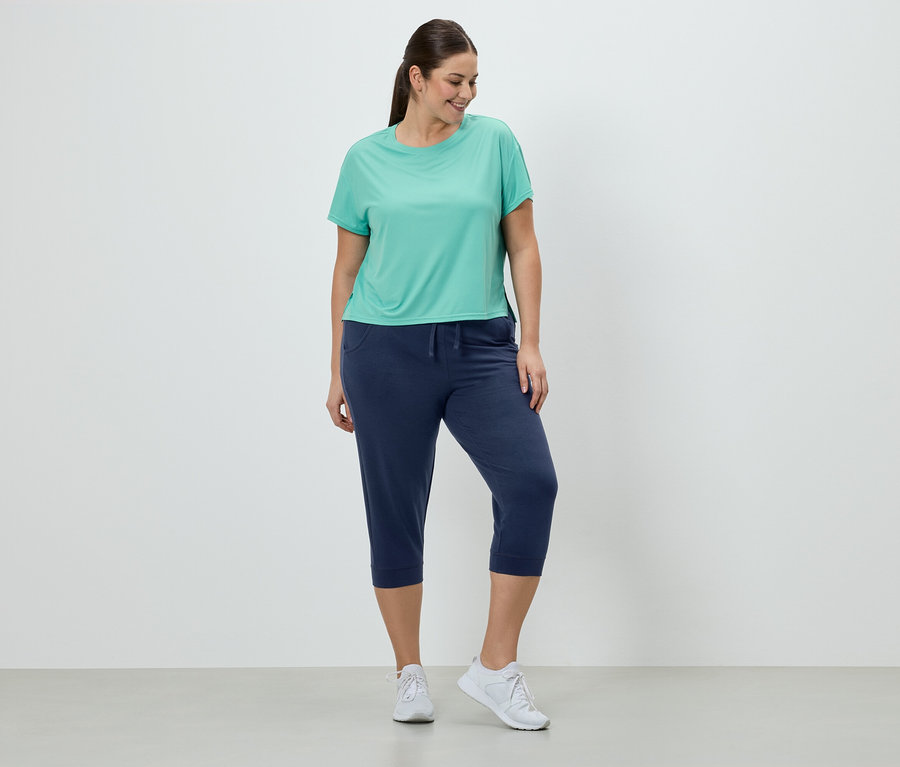 Frau in türkisfarbenem T-Shirt, marineblauer Caprihose und weißen Turnschuhen steht vor einer weißen Wand.