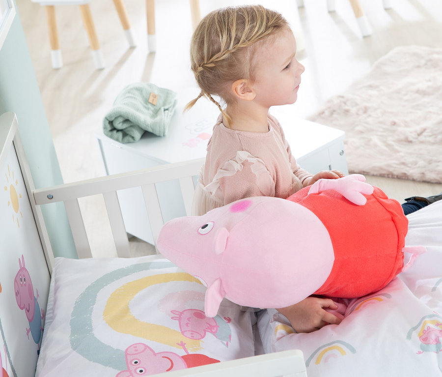 Kleines Mädchen sitzt im Kinderbett mit Peppa Pig Motiv und hält zwei Peppa Pig Stofftiere.