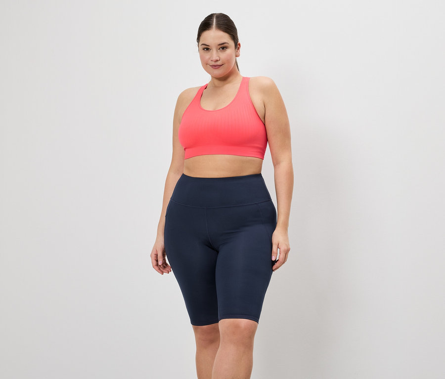 Frau in Sportbekleidung posiert vor einem weißen Hintergrund. Sie trägt einen pinken Sport-BH und kurze, marineblaue Leggings.