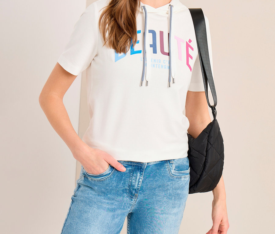 Frau trägt ein weißes CECIL Hoodie-Shirt mit „BEAUTÉ“-Aufschrift, hellblaue Jeans und eine schwarze Stepp-Umhängetasche.