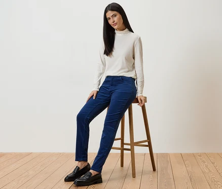 Frau mit langem, dunklem Haar sitzt auf einem Hocker und trägt blaue Cordhose, cremefarbenes Oberteil und schwarze Schuhe.