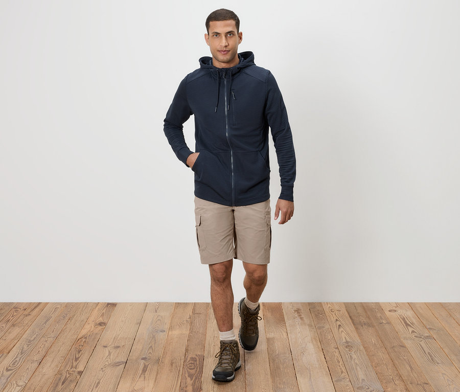 Ein Mann geht in Khaki-Shorts, einem dunkelblauen Kapuzenpullover und braunen Stiefeln auf einem Holzboden.
