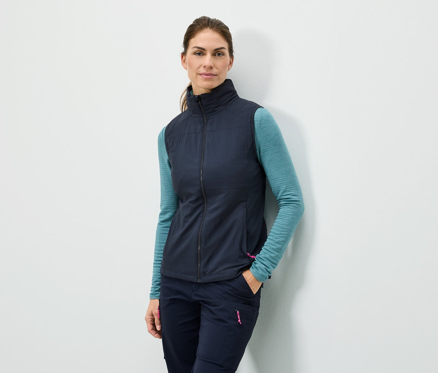 Frau steht mit der Hand in der Tasche an einer Wand, sie trägt eine marineblaue Weste, ein türkisfarbenes Langarmshirt und marineblaue Hosen.