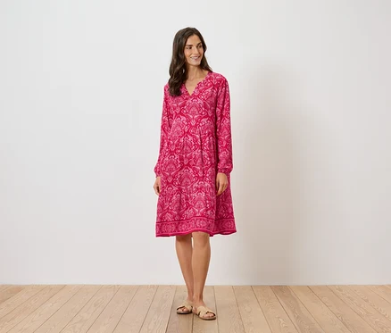 Frau steht und trägt ein pinkes, gemustertes Kleid mit langen Ärmeln und Sandalen.