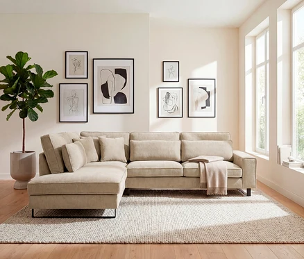 Beige Eckcouch mit Kissen und Decke auf einem hellen Teppich. An der Wand hängen gerahmte Bilder.