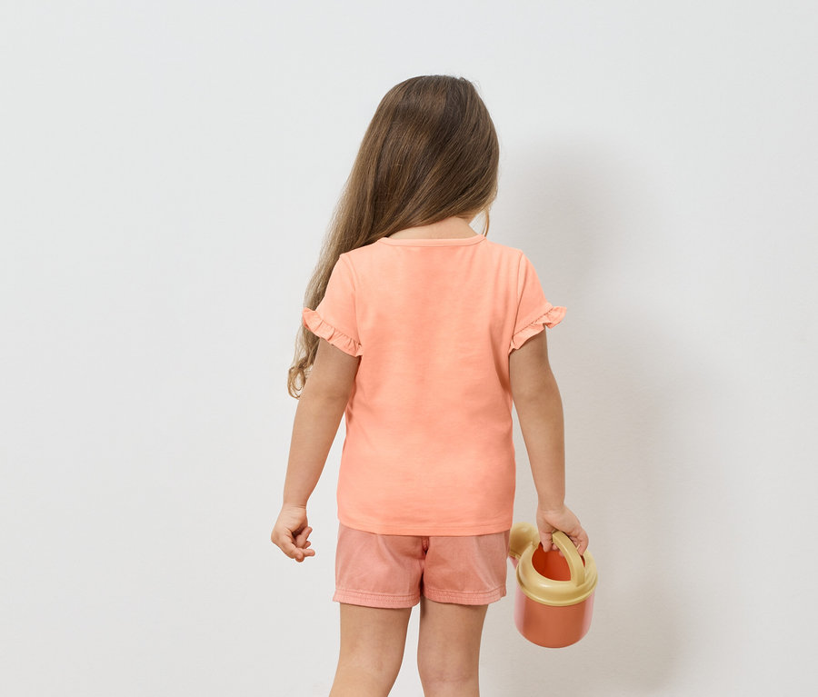 Mädchen von hinten in einem rosa T-Shirt und Shorts, hält eine orangefarbene Gießkanne.