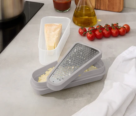 Auf einer Küchentheke stehen ein Topf, Tomatensoße, eine Ölflasche, Tomaten, eine Käsereibe mit Käse und ein Stück Käse in einer Dose.