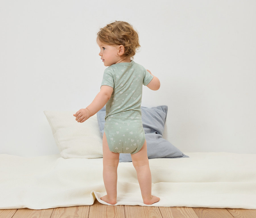 Ein Kleinkind in einem hellgrünen Body mit Herzmuster steht auf einer weißen Krabbeldecke.